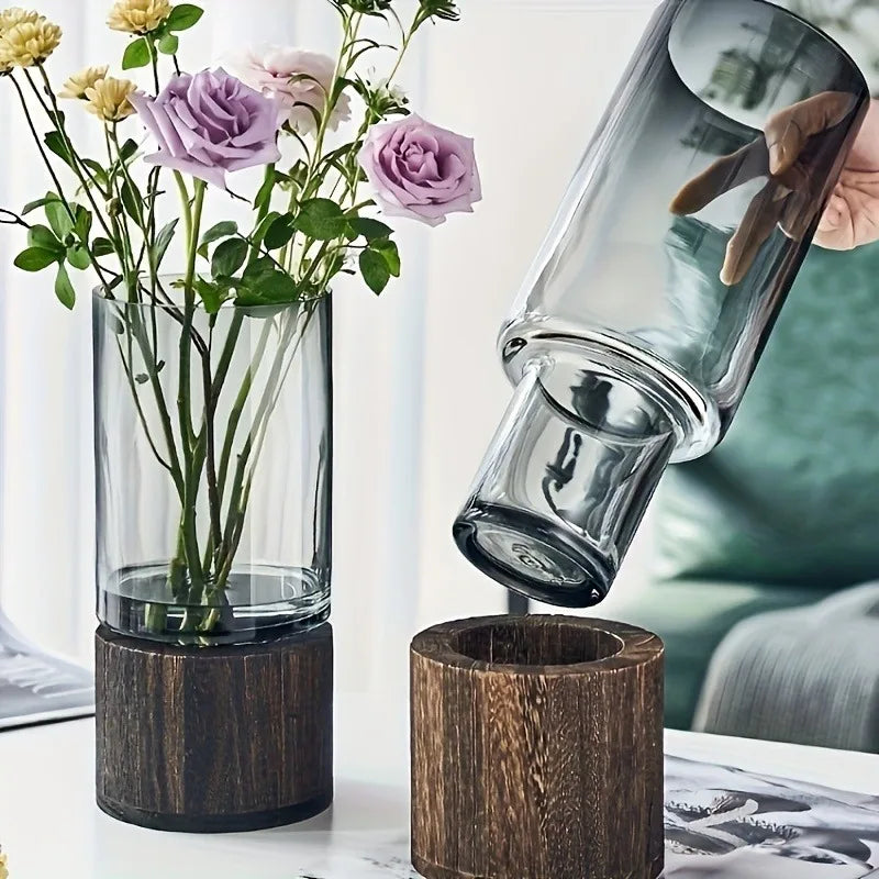 Glass carafe being tilted over a wooden funnel with flowers in a vase on a table.