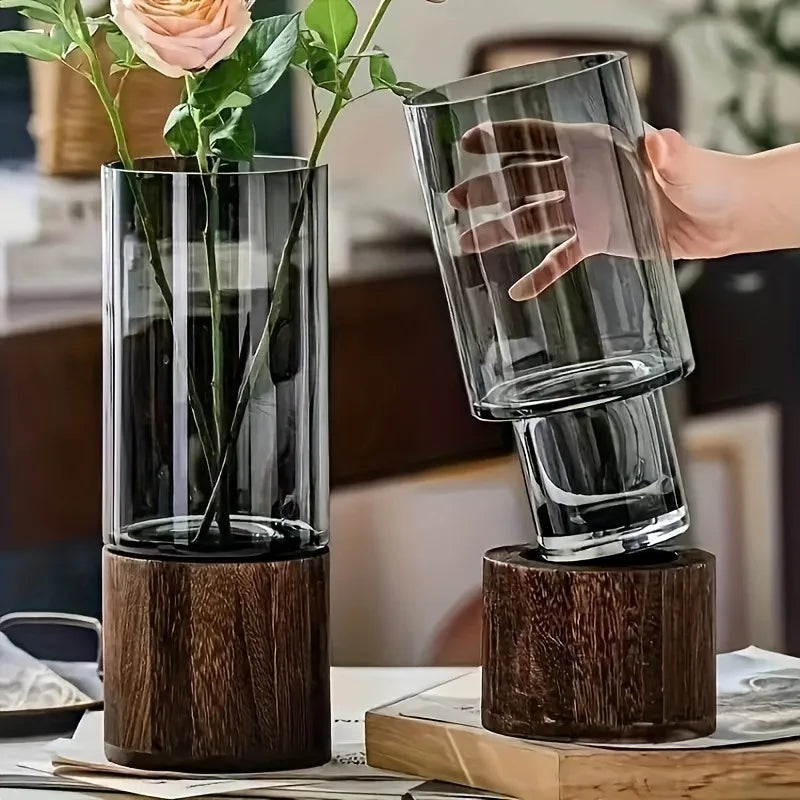 Two glass vases with wooden bases on a table, one being held by a hand.